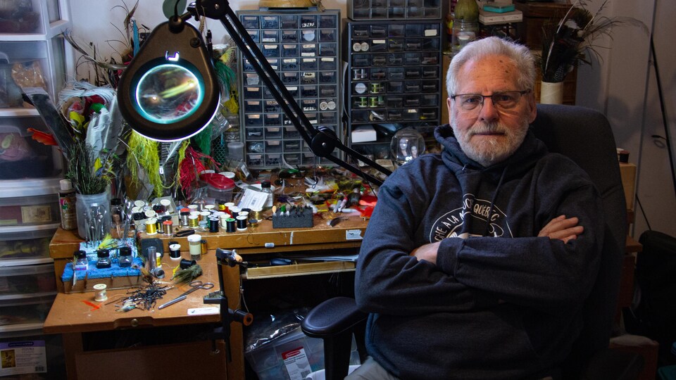 Jacques Turcotte avec les bras croisée regarde la caméra à son bureau sur lequel on y retrouve des plumes, du fils.
