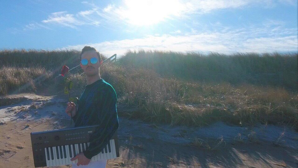 Jalapeño Papa qui porte des lunettes fumées sur une plage avec en main une rose et tenant un clavier sous son bras.
