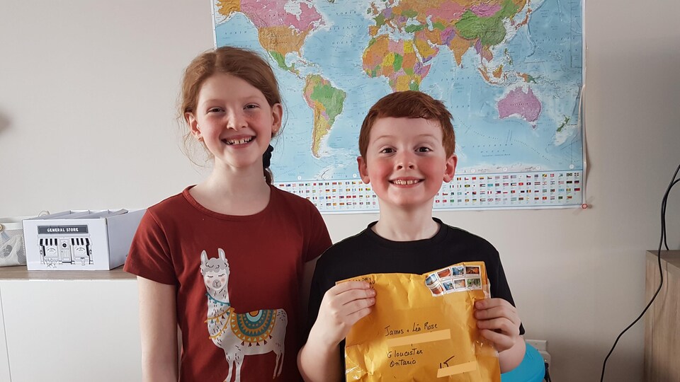 Deux enfants sourient à la caméra. Le garçon tient une enveloppe dans ses mains.