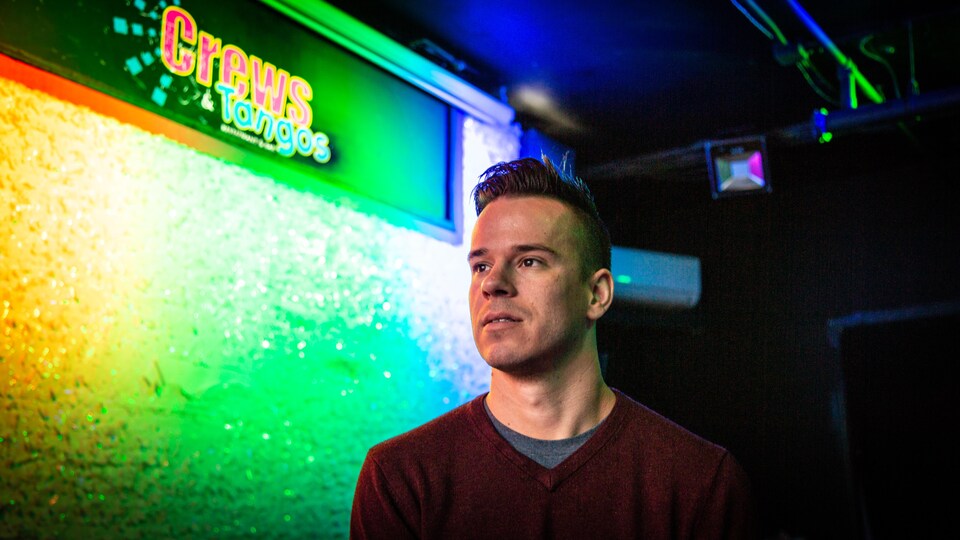 Un jeune homme assis sur une scène devant un mur arc-en-ciel portant le logo du bar Crews and Tango.