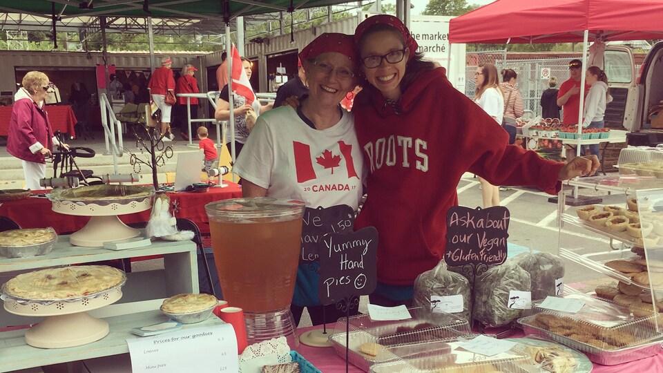 Émilie et Cindy Hébert avec une table garnie de tartes, de gâteaux, de biscuits et d'autres pâtisseries à vendre au marché extérieur