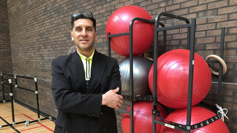 Dans un gymnase, le professeur Taktek se tient debout à côté de ballons d'exercice empilés et rangés. 