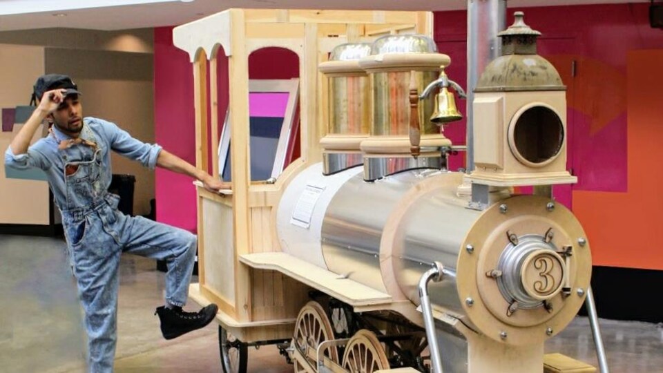 Un homme en habit de conducteur de train, près d'une petite locomotive en bois et en métal.