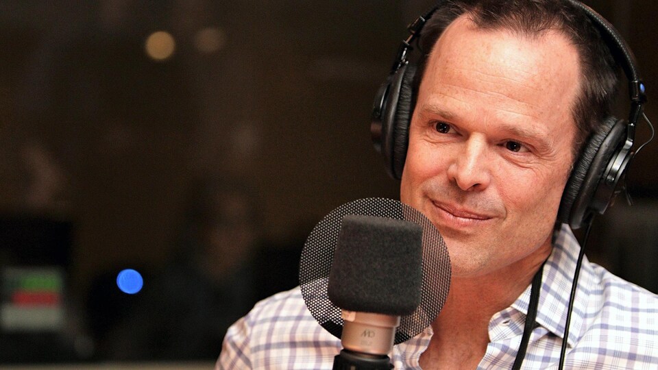 Il affiche un demi sourire devant un micro dans un studio de radio.