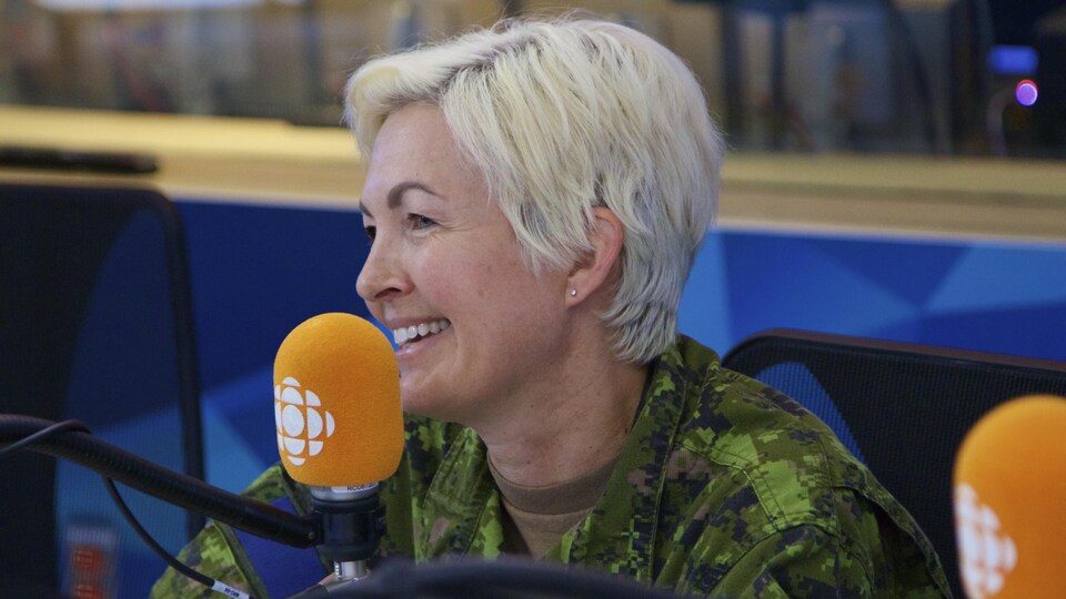 Une femme aux cheveux courts blonds et blancs sourie devant un micro orange dans un studio de radio.