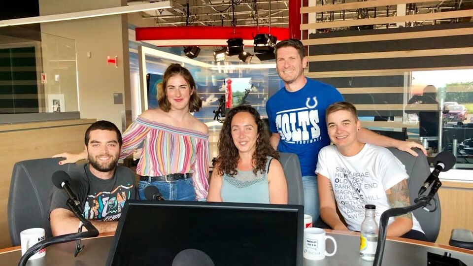 Trois personnes assises et deux personnes debout autour d'une table dans un studio radio.