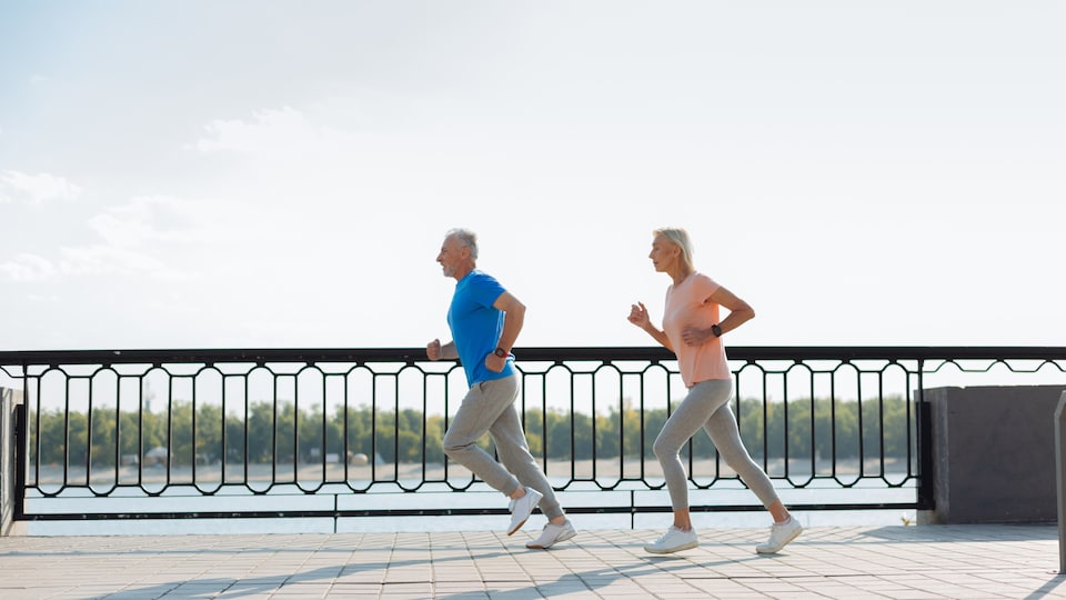 Un couple grisonnant court en vêtements de sports sur une promenade sur le bord de l'eau.