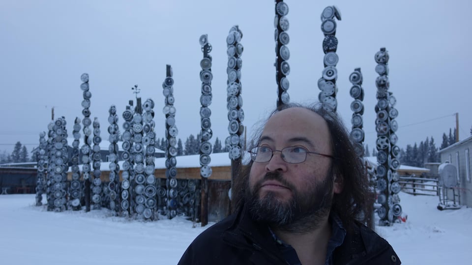 Un homme, le regard vers la gauche, pose devant des sculptures faites avec des jantes de voiture, dehors dans la neige. 