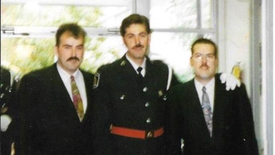 Trois hommes posent côte-à-côte; l'homme au milieu porte un uniforme de policier.