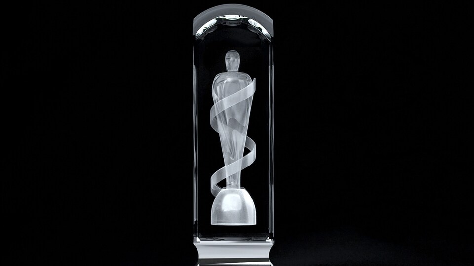 Une statuette rectangulaire en cristal est présentée sur un fond noir. À l'intérieur de ce cristal, est gravé au laser le corps d'une personne qui se tient debout et qui est enveloppé d'un ruban.