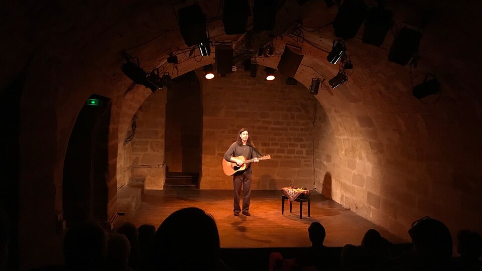 Un musicien seul, sur scène, tenant une guitare et illuminé par des lumières. Derrière lui, un mur de briques. À sa gauche, un tabouret.