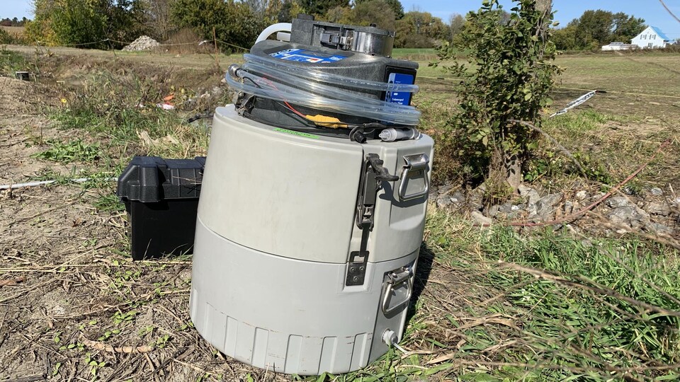 Un appareil scientifique aux allures de gros tonneau gris, muni de boutons de commande et d'un tube, est posé sur un sol agricole.