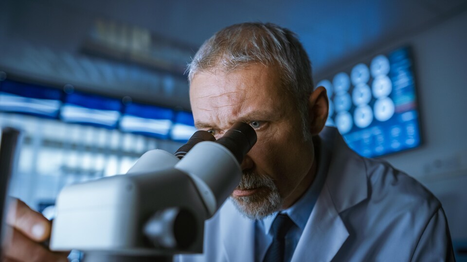 Un homme barbu et grisonnant observe quelque chose à l'aide d'un microscope, dans une grande salle de laboratoire un peu sombre.
