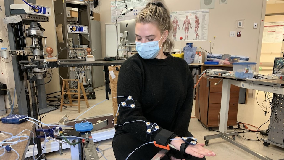 Une jeune femme à la chevelure blonde regarde son bras droit, sur lequel ont été installés des capteurs électroniques, dans un laboratoire de recherche en réadaptation.