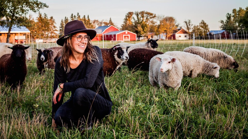 Une jeune femme, portant un chapeau, est accroupie au milieu d'un troupeau de moutons dans un champ.