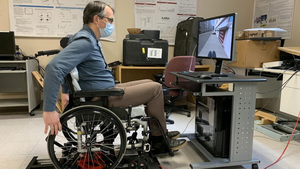 Un homme dans la jeune cinquantaine est assis dans un fauteuil roulant. Devant lui se trouve un écran d'ordinateur sur lequel défilent les images d'une balade sur le trottoir.