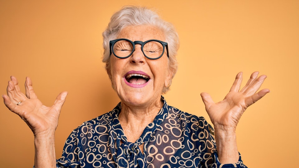 Une dame âgée, aux cheveux gris et portant des lunettes à la mode, s'exclame les yeux fermés, dans la joie.