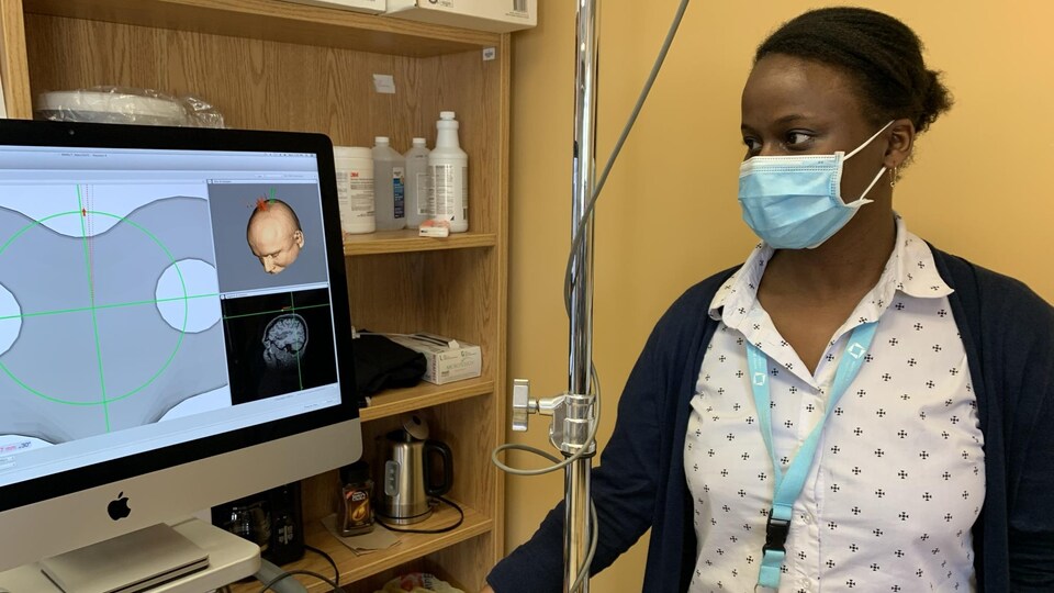 Une femme à la peau noire et aux cheveux court, portant un masque de procédure, regarde un écran d'ordinateur sur lequel apparaît l'illustration d'un crâne.