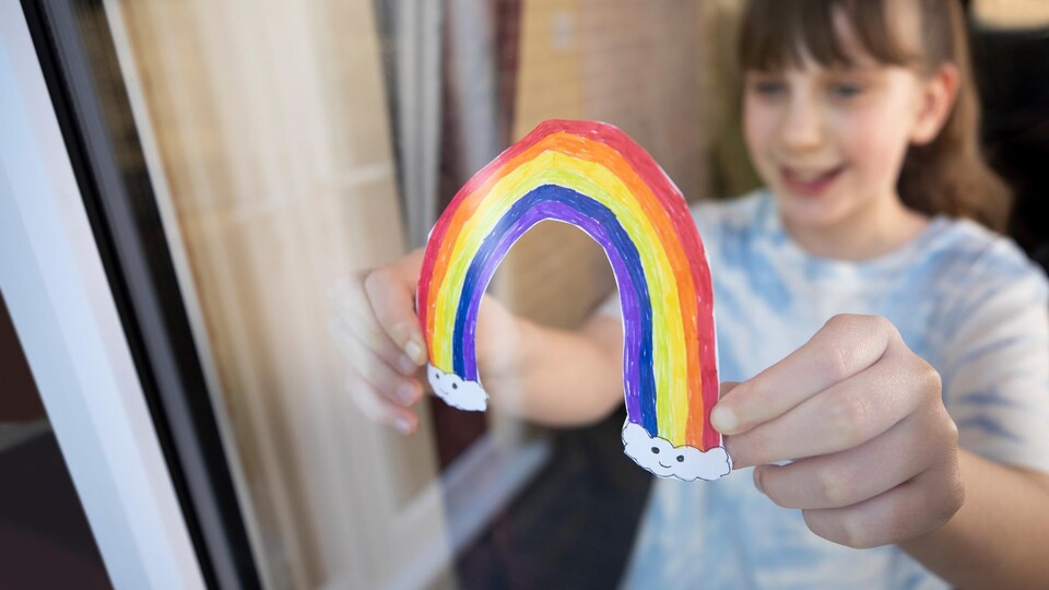 Une fillette fixe un dessin d'arc-en-ciel à une fenêtre.