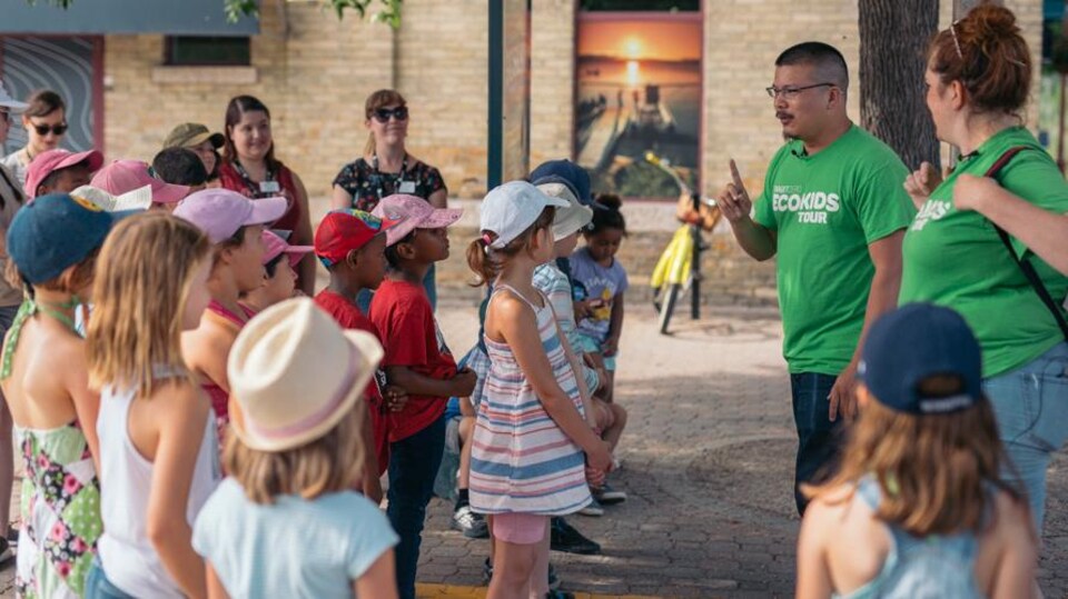 Un comédien vêtu d'un chandail vert, évoquant l'écologie, parle à un groupe d'enfants réunis sous un arbres près d'un édifice public.