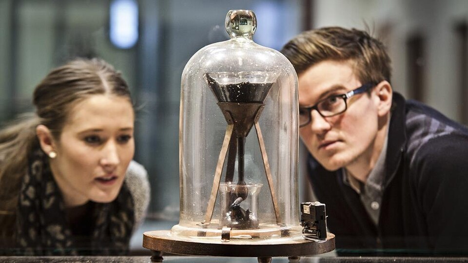 Deux étudiants regardent l'entonnoir contenant la poix sous une cloche de verre.