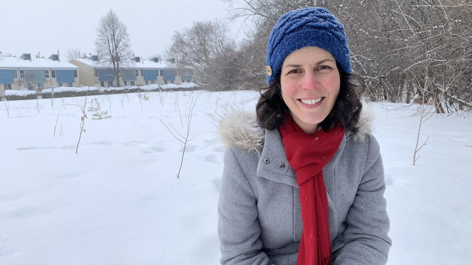 Une jeune femme est accroupie dans un parc couvert de neige, près de petites pousses d'arbres nouvellement plantées. Elle porte un chapeau et un foulard pour se couvrir du froid.