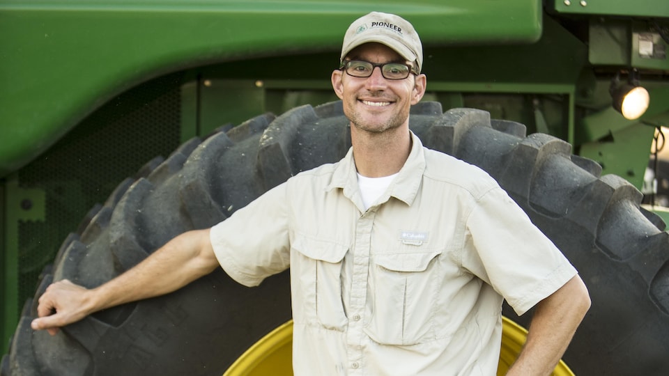 Un homme dans la quarantaine, portant lunettes et casquette, pose fièrement devant un gros tracteur agricole.