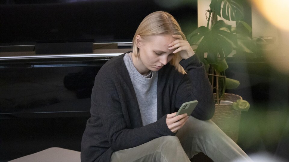 Une jeune femme à la chevelure blonde a les yeux rivés sur son écran de téléphone, assise par terre devant un piano. L'air anxieux, elle semble lire un texte.