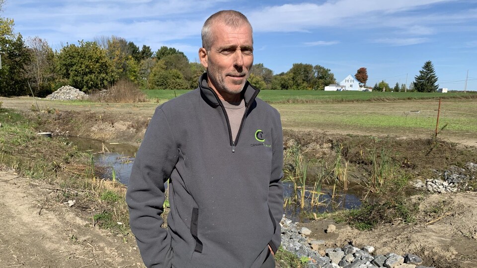 Un agriculteur grisonnant pose pour la caméra, faisant face au soleil, devant un étang d'eau aménagé sur une ferme.