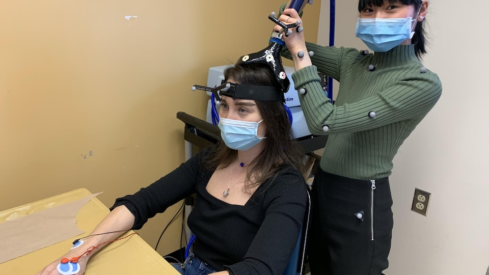 Deux jeunes femmes sont en action dans un laboratoire. L'une est assise et l'autre, debout derrière. La personne assise a un équipement électronique posé sur sa tête.