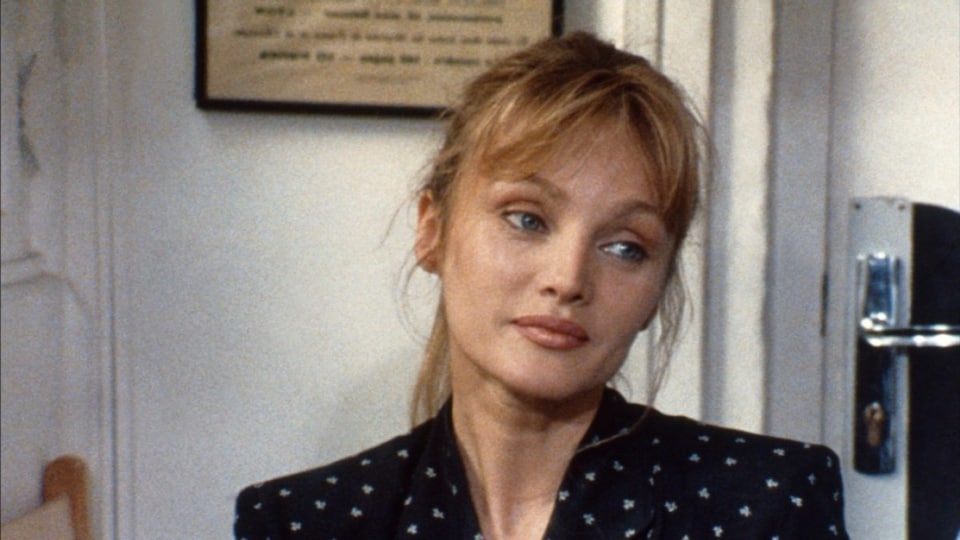 Arielle Dombasle dans une scène du film L'arbre, le maire et la médiathèque (1993), d'Eric Rohmer. Gros plan sur le visage et les épaules de la comédienne aux cheveux blonds et aux yeux bleu perçants, qui porte une chemise foncée à petits motifs blancs et qui regarde son interlocuteur en esquissant un sourire.