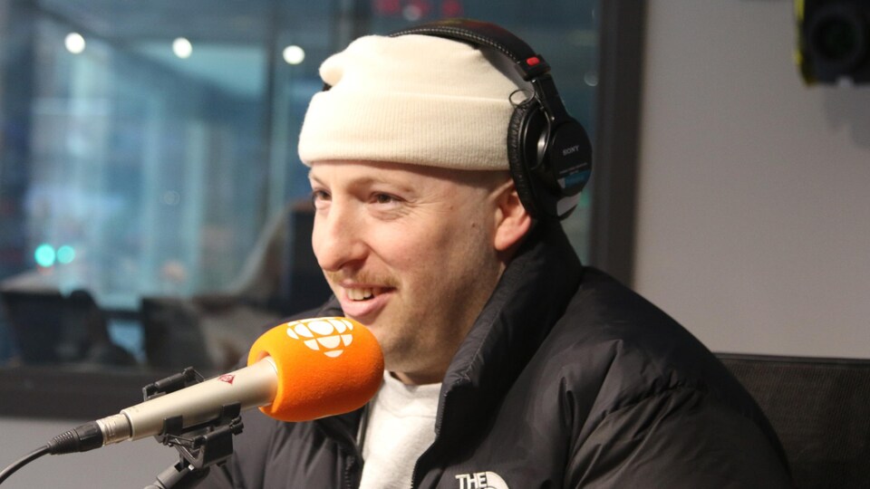 Un homme en veste d'hiver et en bonnet sourit devant le micro.