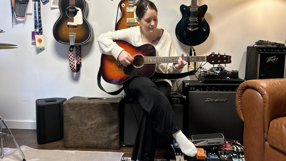 Laura Rae est assise sur un amplificateur dans son studio d'enregistrement et joue de la guitare.