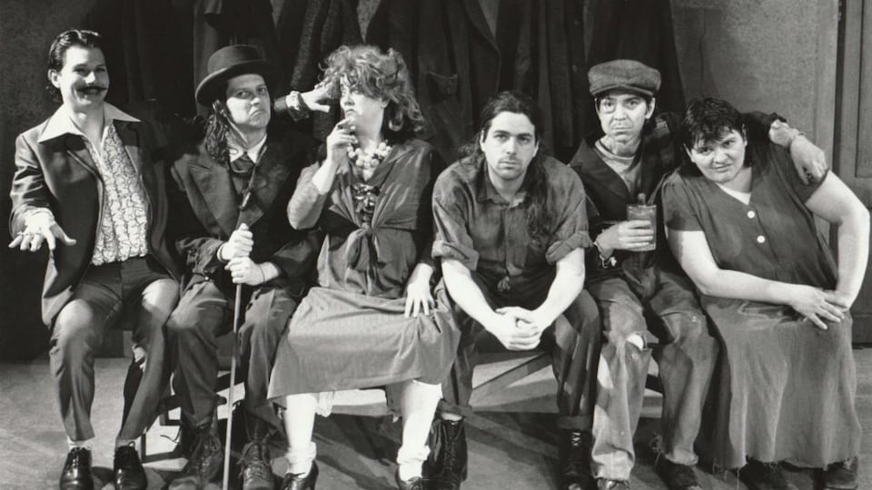 Six hommes et femmes en costumes d’époque sont assis sur un banc.