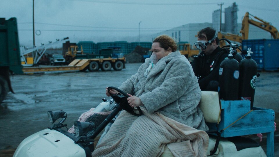 Une femme sur une voiturette de golf, sous des couverture, avec un plongeur et des bonbonne dans une zone industriel.
