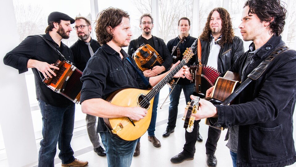 Les membres du groupe Le Vent du Nord jouent de leurs instruments devant de larges fenêtres.
