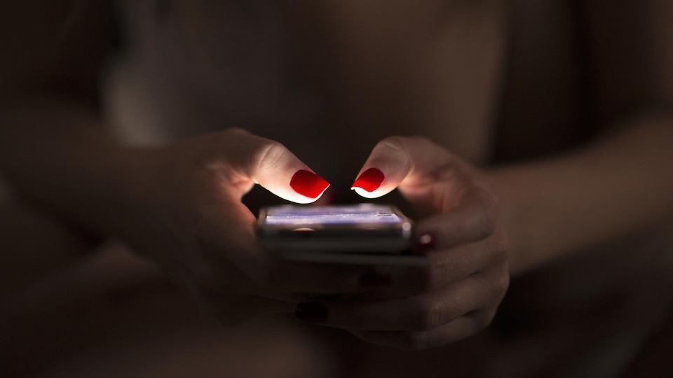 Un gros plan sur les mains d'une femme tenant un téléphone intelligent. Les ongles rouges de la femme sont éclairés par l'écran.