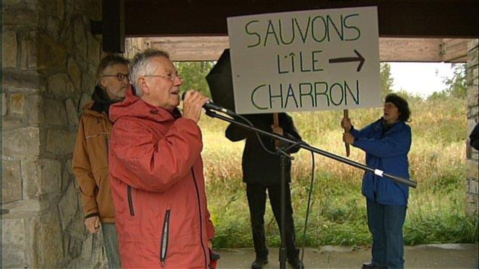 Des personnes manifestent; l'une d'elles tient une pancarte disant «Sauvons l'île Charron».