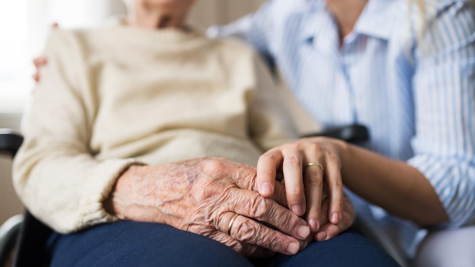 Une personne âgée en fauteuil roulant, à qui une femme plus jeune tient la main.
