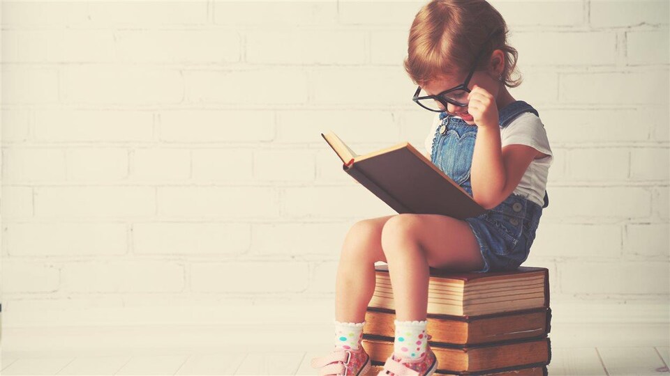 Un enfant lit un livre avec de grosses lunettes, assis sur plusieurs livres.