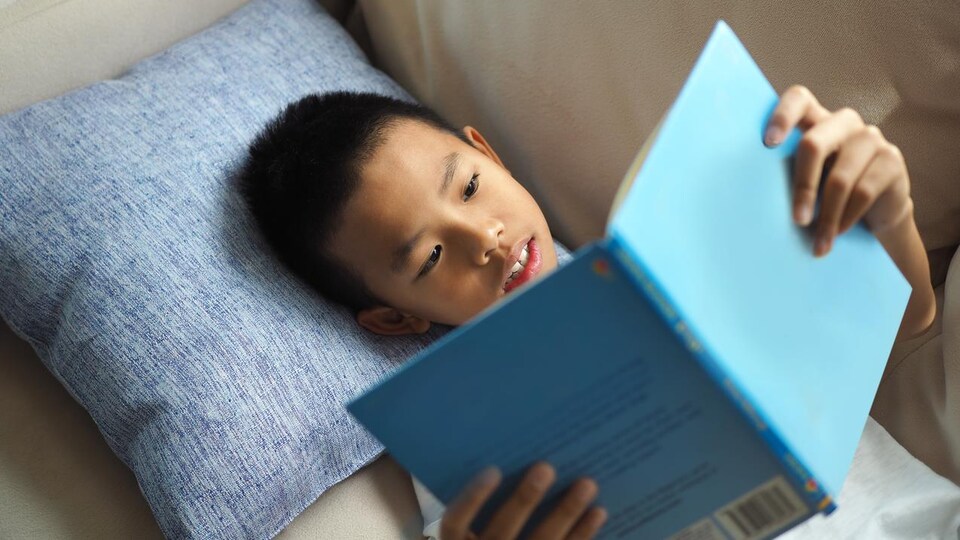 Un jeune garçon lit un livre allongé.