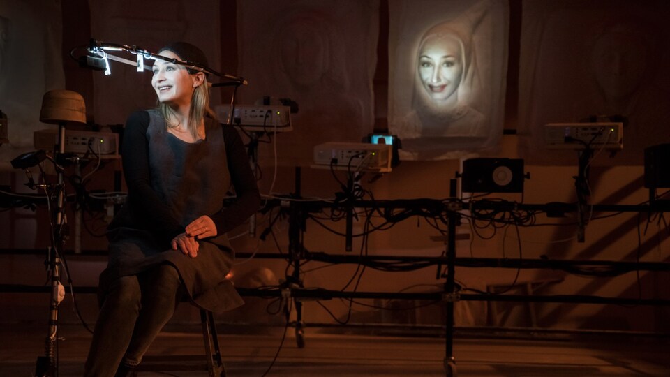 La comédienne porte un casque muni d'une caméra qui filme son visage et le retransmet sous forme de masque vidéo en 3D sur une toile sculptée, derrière elle.