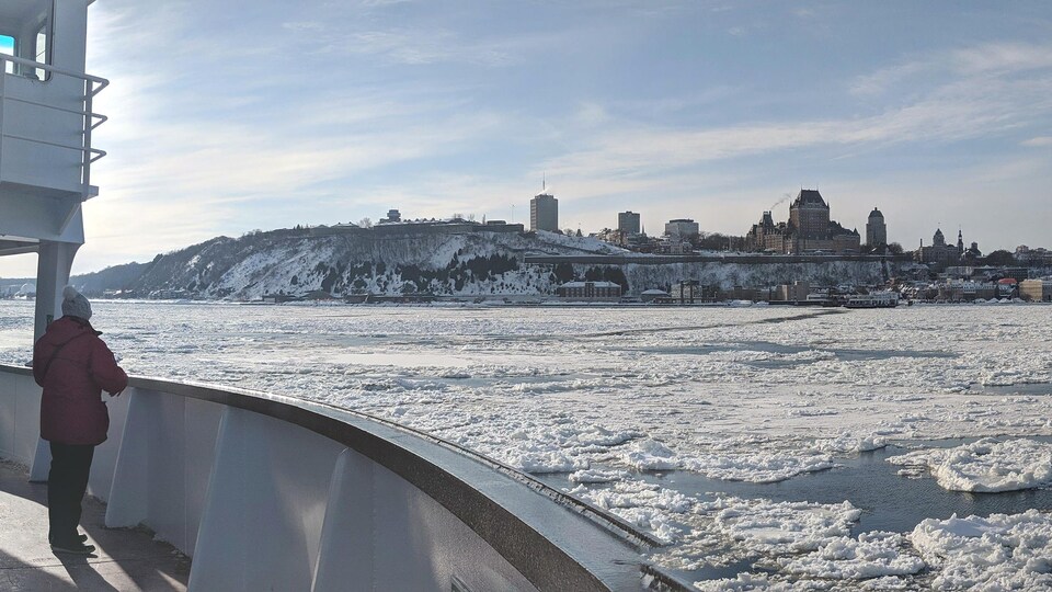 Sur un traversier en hiver sur un fleuve gelé, une personne regarde une ville au loin.