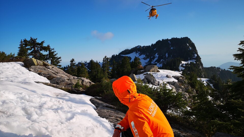 Un homme habillé d'une veste de sauvetage orange. Il est assis dans une montagne en neige et attend l'arrivée d'un hélicoptère