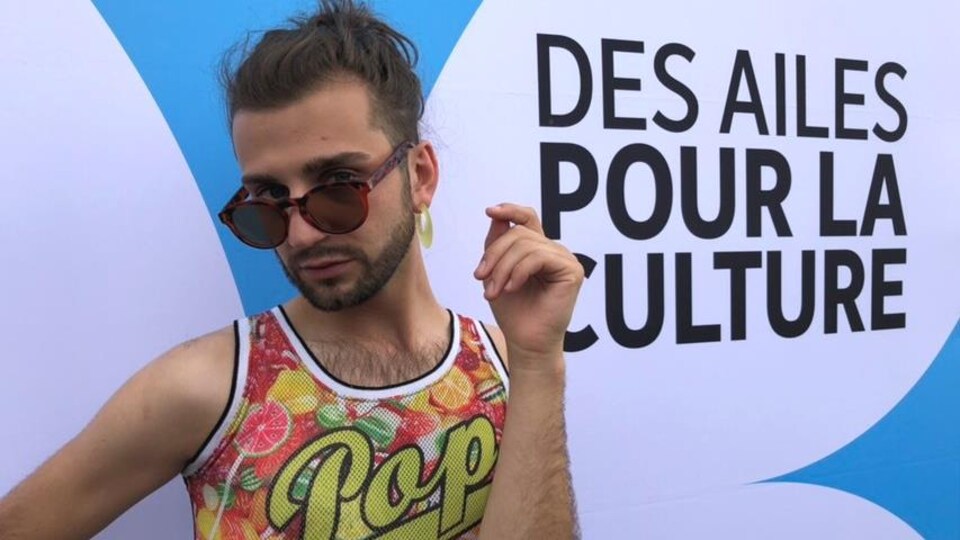 Xavier Gould pose avec des lunettes de soleil devant un panneau avec le libellé « Des ailes pour la culture »