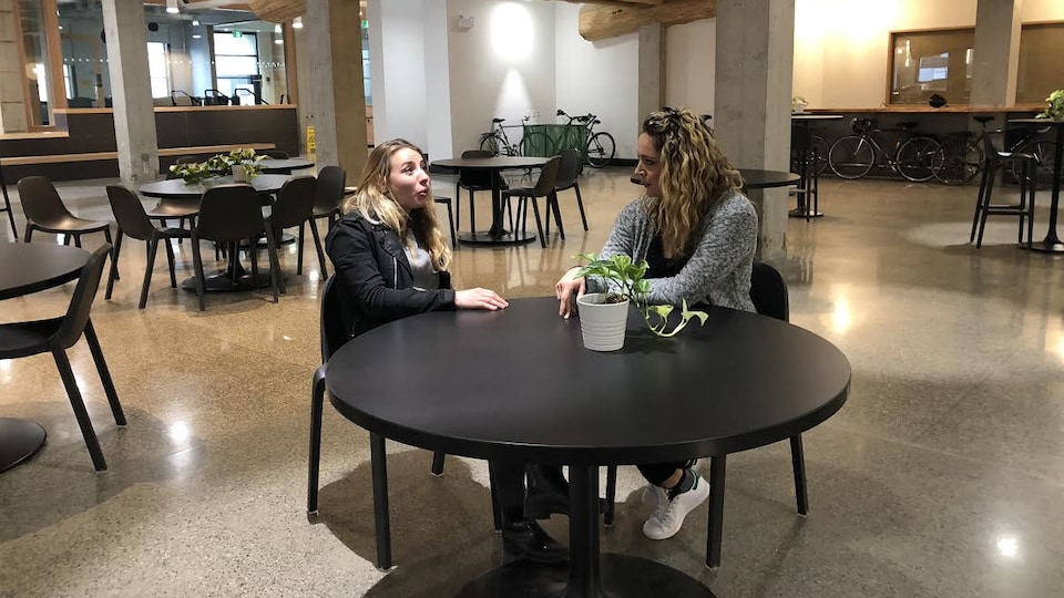 Deux femmes sont assises autour d'une table ronde dans une grande salle vide, et où on voit six autres tables rondes.