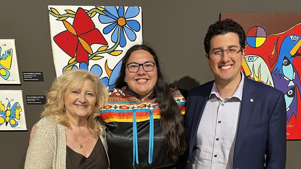 Jasmina Jovanovic (directrice exécutive et conservatrice en chef de la Galerie d'art d'Algoma, Lucia Laford (artiste) et Mark Lepore (président du conseil d'administration) sont photographié devant certaines œuvres de l'exposition Gathering in Honor.