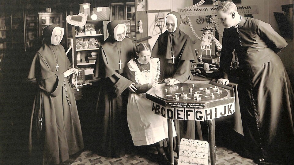 Photo en noir et blanc montrant une jeune fille entourée de trois religieuses et d'un abbé, devant un plateau affichant des lettres de l'alphabet.