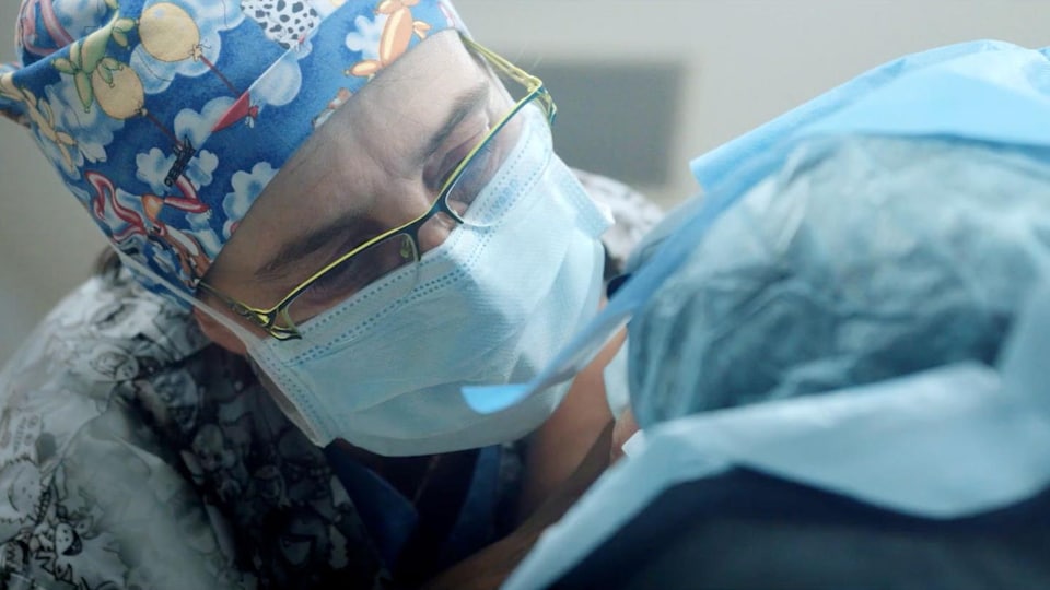 une femme en habit de chirurgien regarde un patient dormir.
