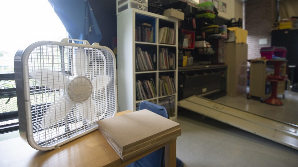 Un ventilateur est posé sur un bureau, devant une fenêtre.